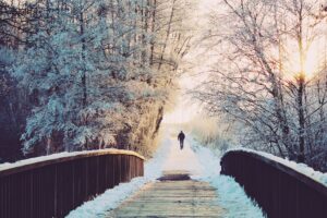 man walking away along snowy track, through woods. A wooden bridge in the foreground. Sunset.